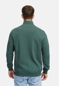 Mand iført en teal ribbet pullover med høj krave og lyseblå jeans, stående med ryggen til mod en ensfarvet baggrund.