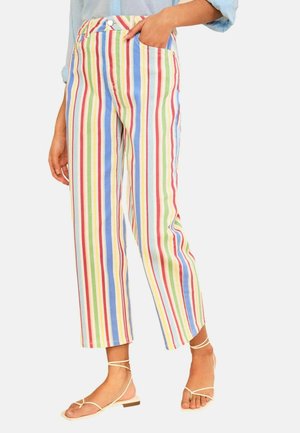 Osoba nosiaca farebné vertikálne pruhované nohavice s vysokým pásom a skrátenými nohavicami, biele ploché sandále so remienkami, ruka v vrecku.