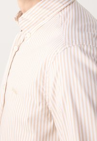 Chemise beige et blanche à rayures verticales boutonnée portée par une personne, montrant le col, la couture de l'épaule et une partie de la poitrine.