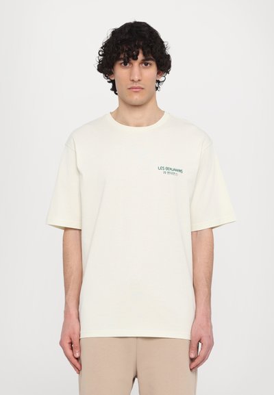 T-shirt en coton couleur crème avec une coupe décontractée, manches courtes et texte vert sur la poitrine gauche. Design minimaliste, texture lisse.