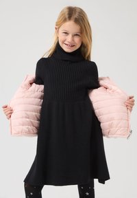 Abito nero a coste con collo alto, abbinato a una giacca morbida rosa chiaro tenuta aperta. Tessuto strutturato; leggings scuri con motivo a stelle.