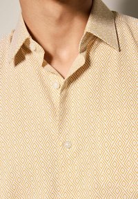 Camisa amarilla de botones con un patrón de diamantes repetido en blanco. La tela tiene una textura suave, con un cuello clásico y una tapeta de botones.
