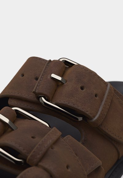 Κοντινό πλάνο καφέ σουέντ λουριών σανδαλιών με ασημένιες μεταλλικές αγκράφες και πολλαπλές τρύπες ρύθμισης.