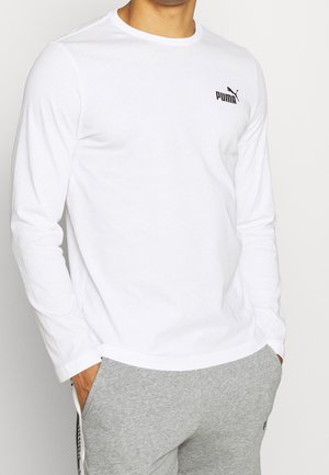 Mann trägt ein weißes Puma Langarmshirt und graue Jogginghosen, Hände in den Taschen, vor einem schlichten Hintergrund.