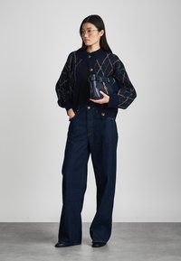 Cardigã com padrão marinho e mangas bufantes, com fecho de botão. Top preto, jeans largos escuros e uma mala azul-marinho com alça. Óculos transparentes.