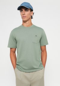 Jonge man in een lichtgroene t-shirt met een voorzak en een klein logo, beige broek en een blauwe honkbalpet, staand met zijn handen in de zakken.