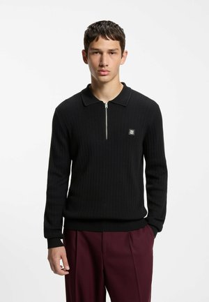 Ung mand med alvorligt udtryk iført en sort lynlås sweater og bordeaux bukser foran en ensfarvet hvid baggrund.