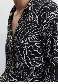 Chemise noire arborant des motifs floraux et de feuilles blancs, avec un col et une poche poitrine. Fabriquée en tissu léger à texture lisse.