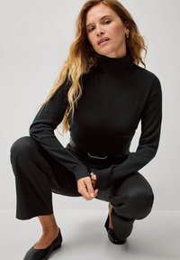 Maglione nero con collo alto e maniche lunghe, realizzato in un tessuto morbido. Abbinato a pantaloni neri a vita alta e scarpe basse.