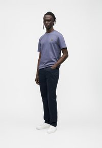 Camiseta de algodón morada con un pequeño logo amarillo, combinada con pantalones azul oscuro y zapatillas blancas. El modelo posa frente a un fondo liso.