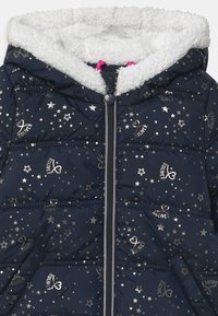 Veste matelassée marine avec des motifs d'étoiles argentées et de cœurs, capuche doublée en fausse fourrure, et tirette rose, présentant un design matelassé et des accents de logo.