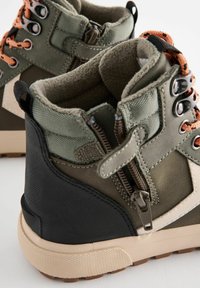 Scarponi da trekking verdi e neri con design alto. Le caratteristiche includono una cerniera laterale, tessuto testurizzato e lacci arancioni. Suola in gomma marrone.