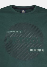 Zelené bavlněné tričko s kulatým grafickým motivem a tmavými pruhy, na kterém je text "WRECKING CREW PETROL ALASKA" v kontrastních barvách.