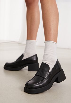 Zwarte leren loafers met een ronde neus, stevige blokhak en een decoratieve band over de wreef, gedragen met witte enkelkousen.