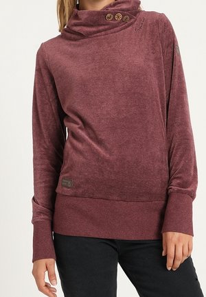 Woman wearing a long-sleeve, maroon cowl neck sweater with three wooden buttons on the collar, paired with black pants.