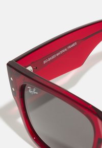 Ochelari de soare roșii cu rame din material bio, având un design elegant, lentile gri și un logo notabil Ray-Ban pe braț.