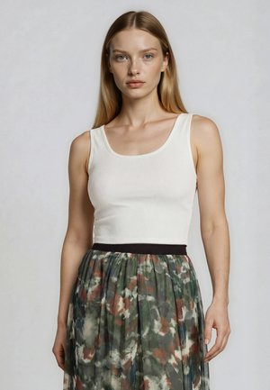 Vrouw met lang blond haar draagt een witte mouwloze top en een geplooide rok met hoge taille met een groen, bruin en grijs abstract patroon.