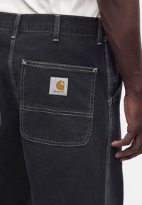 Dunkelgraue Jeans mit weißen Nähten, mit einer quadratischen Gesäßtasche mit einem Carhartt-Logo-Patch, getragen von einer Person mit dunkler Haut.