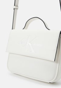 Bolso de hombro de imitación cuero blanco con ribete negro, que presenta un cierre de solapa y un logotipo "CK" grabado en la parte frontal. Textura suave, forma compacta.