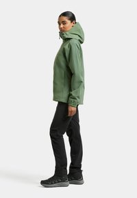 Grüne wasserdichte Jacke mit Kapuze, elastischen Bündchen und Seitentaschen, kombiniert mit schwarzer Hose und robusten schwarzen Outdoor-Schuhen.
