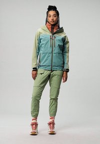 Giacca leggera in tre tonalità, turchese, verde e beige, con tasche con zip, abbinata a pantaloni verdi e scarpe multitonali.