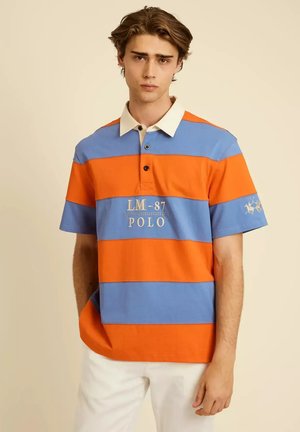 Jonge man draagt een blauw-oranje gestreept poloshirt met de tekst "LM-87 POLO", een witte kraag en witte broek, staand tegen een beige achtergrond.