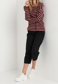 Gestreiftes Langarmshirt in Burgunderrot und Hellrosa, kombiniert mit schwarzen kurzen Hosen und weißen Sneakers, vor einem einfarbigen Hintergrund.