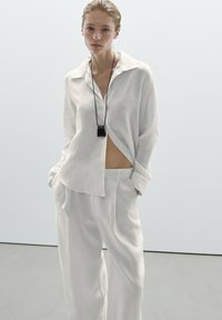 Bílá lněná košile s volným střihem, s špičatým límcem a předními knoflíky, spárovaná s volnými kalhotami ve stejném stylu. Černý náhrdelník s přívěskem.