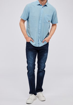 Mężczyzna w jasnoniebieskiej koszuli z krótkim rękawem na guziki, ciemnoniebieskich dżinsach i białych trampkach, stojący z rękami w kieszeniach.