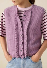 Femme portant un gilet tricoté lavande avec des bords en dentelle sur un t-shirt rayé rose et bordeaux et un jean bleu clair.