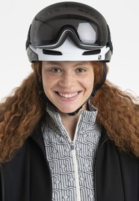 Skihorn in Weiß mit einem großen dunklen Visier, getragen über einer gemusterten Zip-Up-Jacke mit grauen und schwarzen Akzenten. Sommersprossen im Gesicht sichtbar.