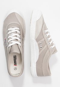 Beige canvas sneakers with white rubber toe cap and textured sole. Features white laces, metallic eyelets, and a logo on the side.