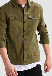 Veste en denim vert olive avec fermeture à boutons, deux poches poitrine et un ourlet droit. Le tissu est légèrement texturé.