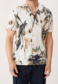 Camicia a maniche corte con bottoni di colore bianco, decorata con un motivo a foglie di palma nei toni del verde, marrone e blu scuro. Presenta una texture morbida e una vestibilità rilassata.