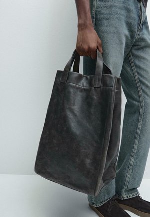 Borsa a tote nera in pelle con manici robusti, forma rettangolare, texture liscia e design minimalista, tenuta da una persona in denim.