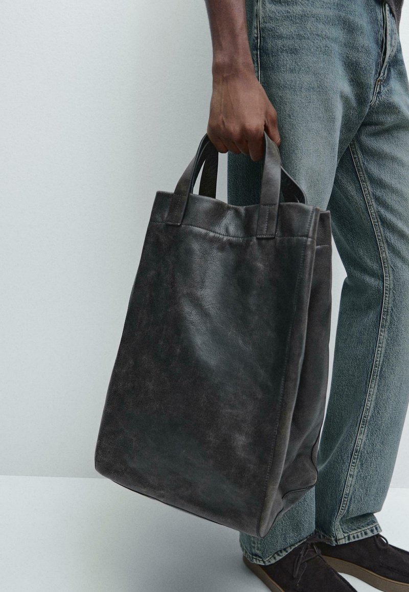 Zwarte leren tote bag met stevige handvatten, rechthoekige vorm, gladde textuur en een minimalistisch design, vastgehouden door een persoon in denim.