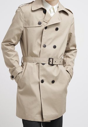 Trench-coat beige croisé avec des boutons noirs et une ceinture, porté sur une chemise blanche, mains dans les poches, sur un fond uni.