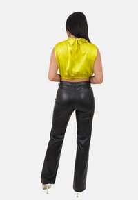 Kobieta z długimi ciemnymi włosami, ubrana w bezrękawnik ze jasnego, żółtego satynu związanego na szyi, czarne skórzane spodnie i białe szpilki.