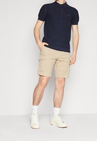 Homme portant un polo en maille navy, un short cargo beige, des chaussettes blanches et des baskets crème, se tenant devant un fond blanc.