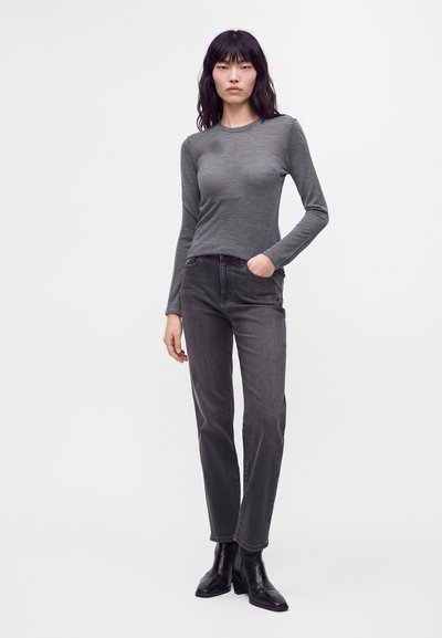 Femme aux longs cheveux noirs porte un haut gris ajusté à manches longues, un jean gris foncé et des bottines noires, debout devant un fond blanc uni.
