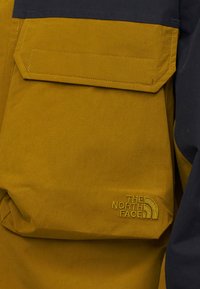 Κοντινό πλάνο τσέπης μουσταρδί μπουφάν με καπάκι και μαύρο επάνω τμήμα, με κεντημένο λογότυπο "The North Face" σε αντίστοιχο κίτρινο νήμα.
