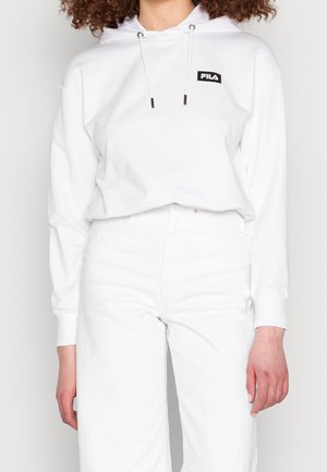 Persoon draagt een witte FILA hoodie met trekkoorden en witte hoge taillerokken, staand tegen een effen achtergrond.