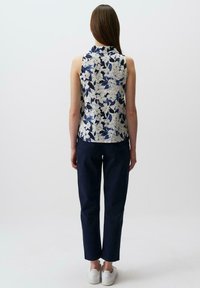Kwiatowy top bez rękawów w niebiesko-kremowym wzorze, zestawiony z granatowymi spodniami. Modelka stoi tyłem, nosząc białe trampki.