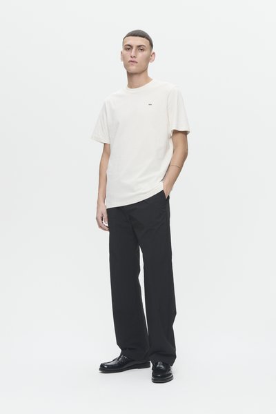 Lichtcrème katoenen t-shirt met korte mouwen en een ronde hals, gecombineerd met losse zwarte broeken en glanzende zwarte leren schoenen.