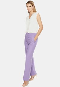 Biała bezrękawnikowa bluzka z dekoltem w serek, zestawiona z jasnofioletowymi spodniami o dopasowanym kroju. Modelka stoi z rękami w kieszeniach, w cielistych szpilkach.