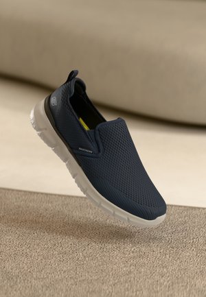 Granatowy but typu slip-on marki Skechers z białą podeszwą na beżowej, fakturowanej powierzchni, prezentujący oddychającą siateczkową cholewkę i detale z logo.