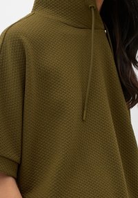 Sudadera verde oliva con un patrón texturizado, cuello de media cremallera y un corte holgado. Cuenta con mangas cortas y un ligero brillo en la tela.