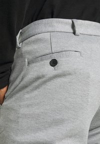 Szare, tkane spodnie o gładkiej fakturze, z tylną kieszenią zapinaną na czarny guzik. Pas jest elastyczny z szlufkami na pasek.