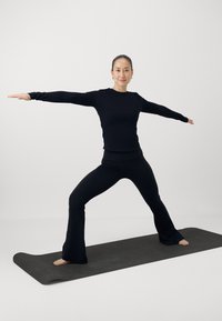 Top nero a maniche lunghe e pantaloni yoga svasati indossati da una persona in posizione guerriero su un tappetino yoga grigio, su uno sfondo bianco semplice.