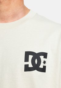 Cremefarbiges Baumwoll-T-Shirt mit einem auffälligen schwarzen DC-Logo und einem Stern. Rundhalsausschnitt und weiche Textur. Einfaches Design, ohne zusätzliche Muster.